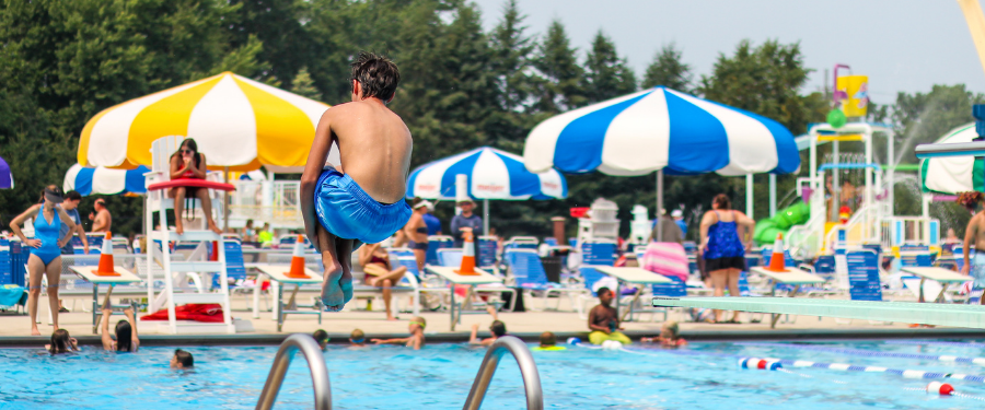 A boy jumps into a pool of people swimming while a lifeguard looks on