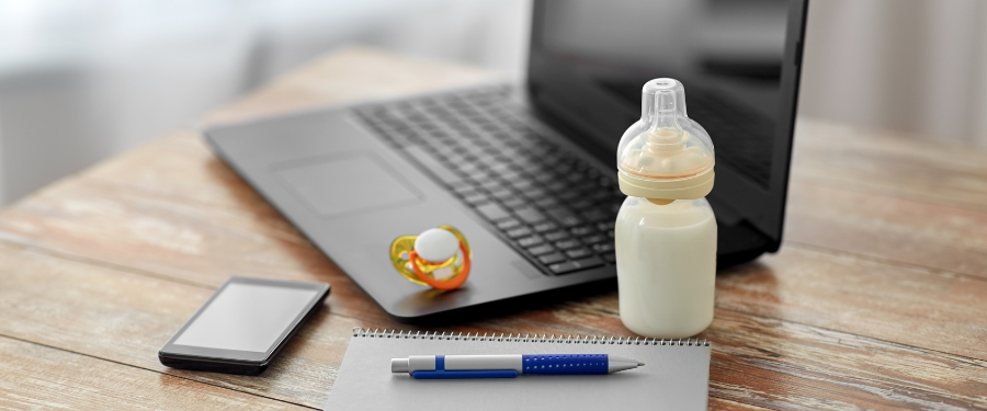 Laptop, milk bottle, pen, notepad, and baby pacifier sitting on a desk 