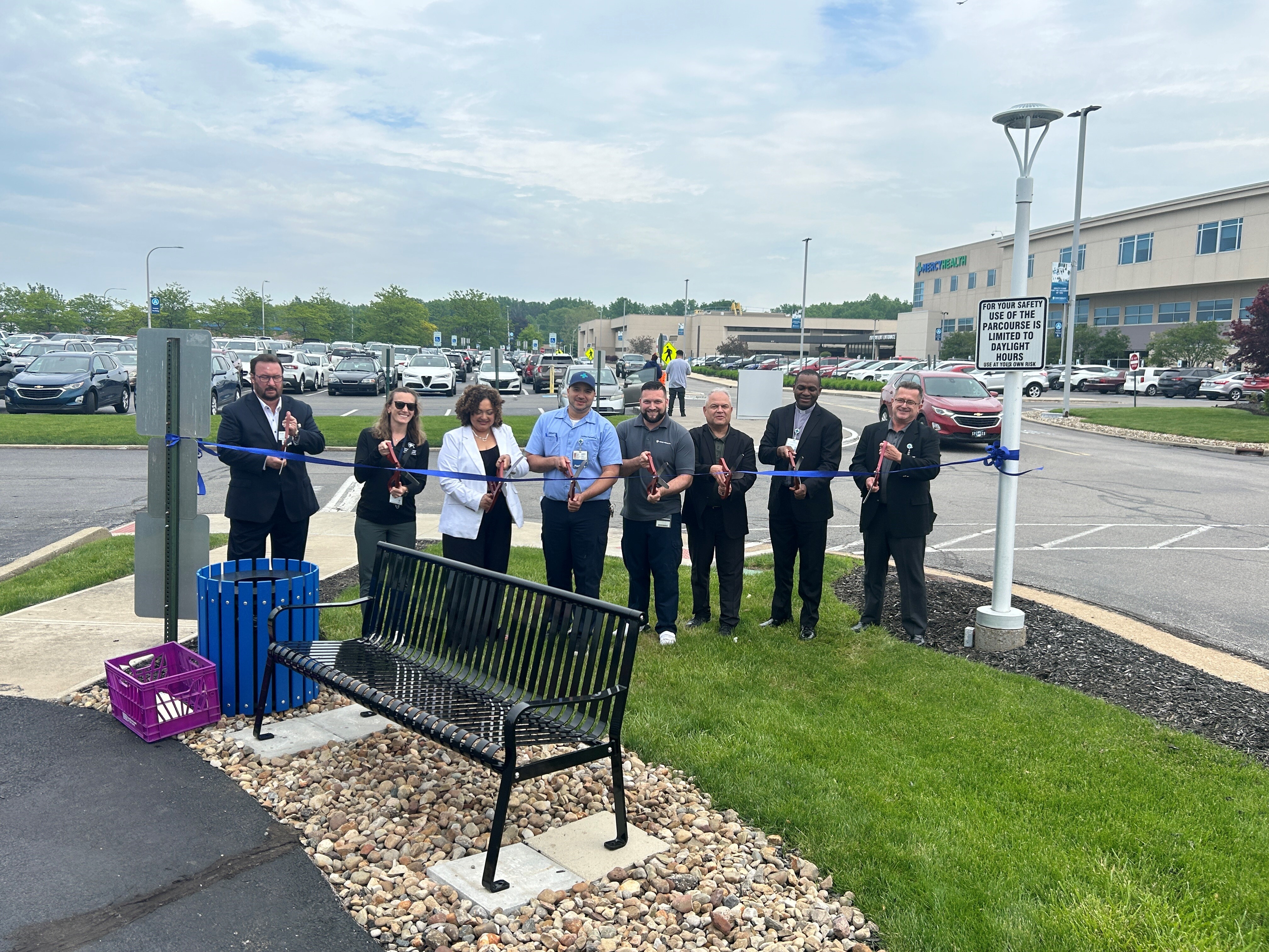 8 people cutting a blue ribbon to unveil a black bench and blue trash receptacle